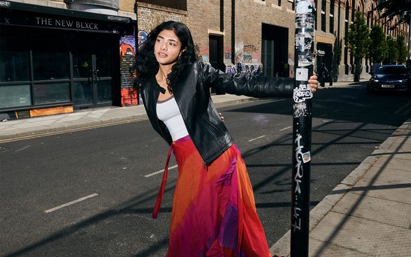 Young woman on a city pavement holding a lamppost, wearing a black leather jacket, white top, and red-orange maxi skirt.