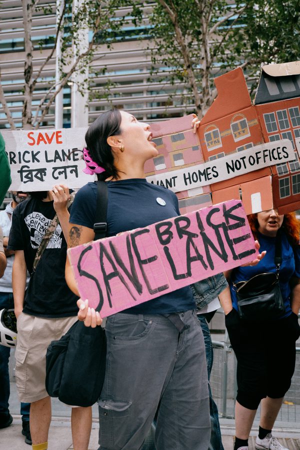 Protester shouting, holding a pink "Save Brick Lane" sign. Crowd behind with banners reading "Save Brick Lane" in Bengali and "Homes Not Offices".