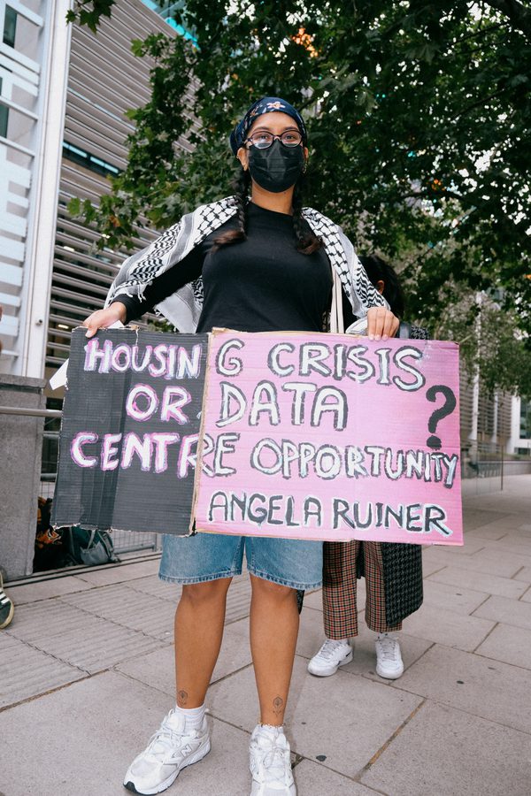 Protestor wearing a black mask and keffiyeh holds signs reading "Housing Crisis or Data Centre Opportunity? Angela Ruiner" on a city pavement.