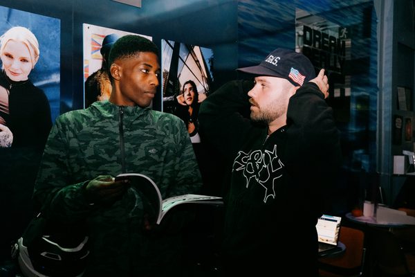 Two men talking indoors near large printed photos on a wall; one in a camo jacket, the other in a black hoodie and cap.