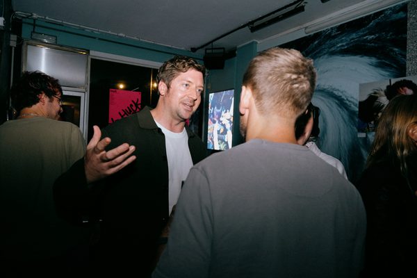 Two men talking at an indoor event or gallery opening, with artwork visible on the walls behind them and other guests in the background.