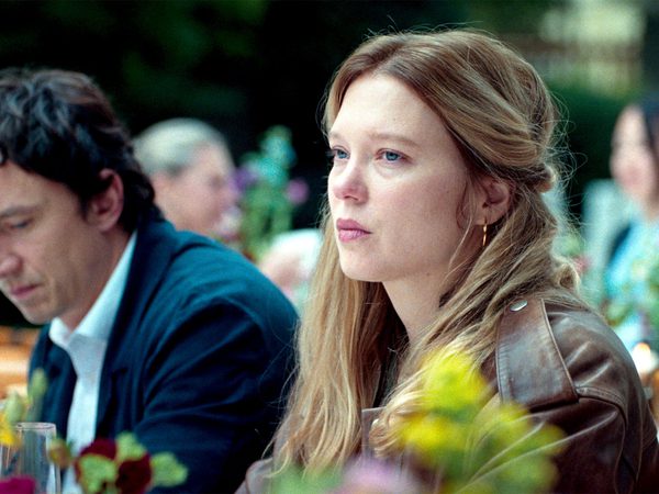 Young woman with light brown hair in a half-up style, wearing a brown leather jacket, seated outdoors at a table with flowers, other guests blurred behind her.