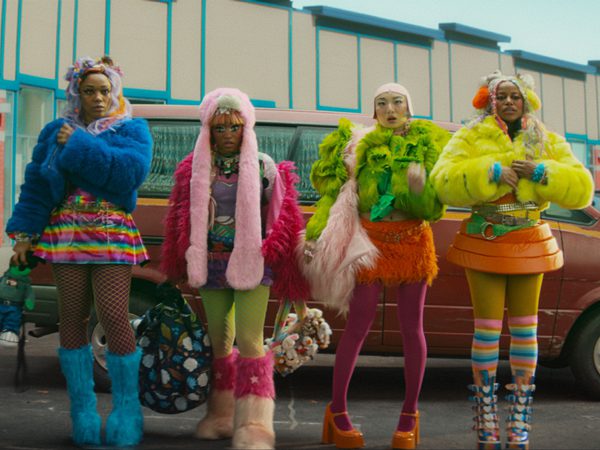 Four people in bold, colourful fluffy outfits standing in front of a vintage car outside a pastel-coloured building.