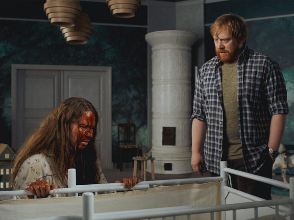 A bloodied woman leans over a white cot as a bearded man in a plaid shirt looks on, in a teal-walled room with a white tiled stove.