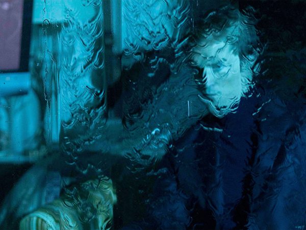 Two people seen through a rain-covered window, bathed in blue light. One person wearing glasses is visible on the right side.