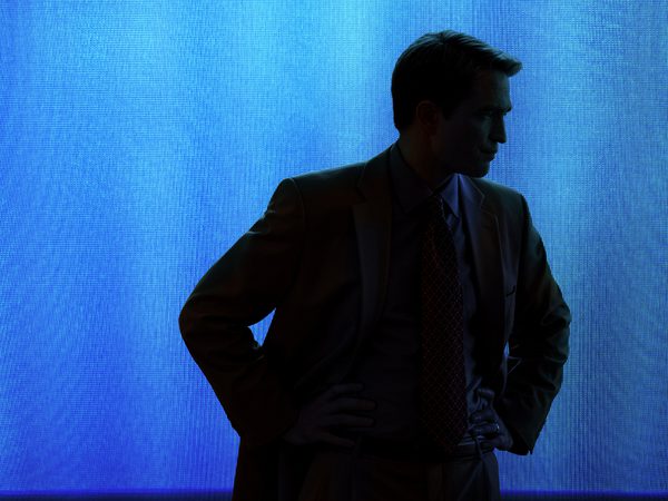 Man in a suit standing with hands on hips, shown in dark silhouette against a bright blue illuminated background.